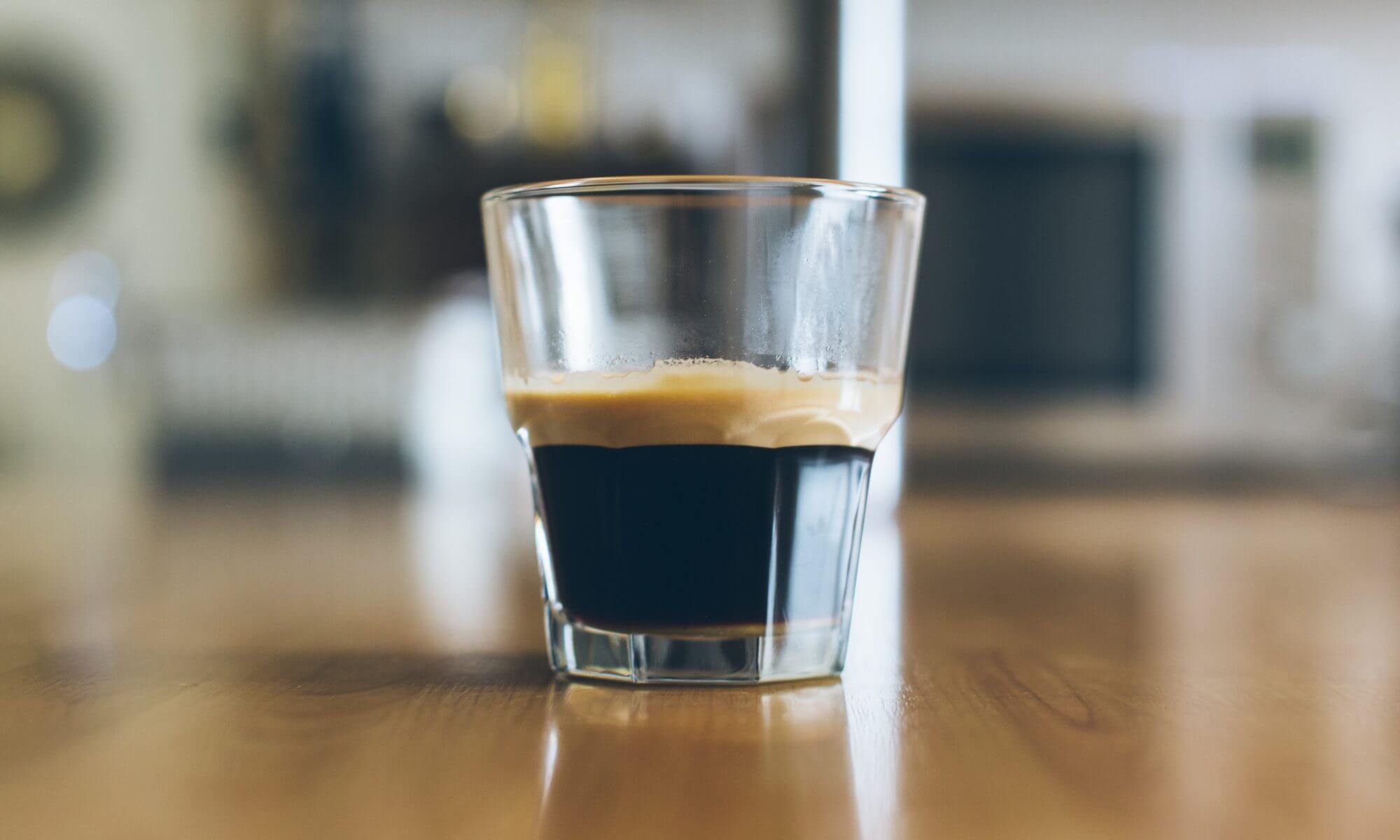 A small glass of dark espresso coffee with a thin layer of foam resting on a warm wooden tabletop, with a softly blurred interior in the background and gentle ambient light in earthy brown and cream tones.