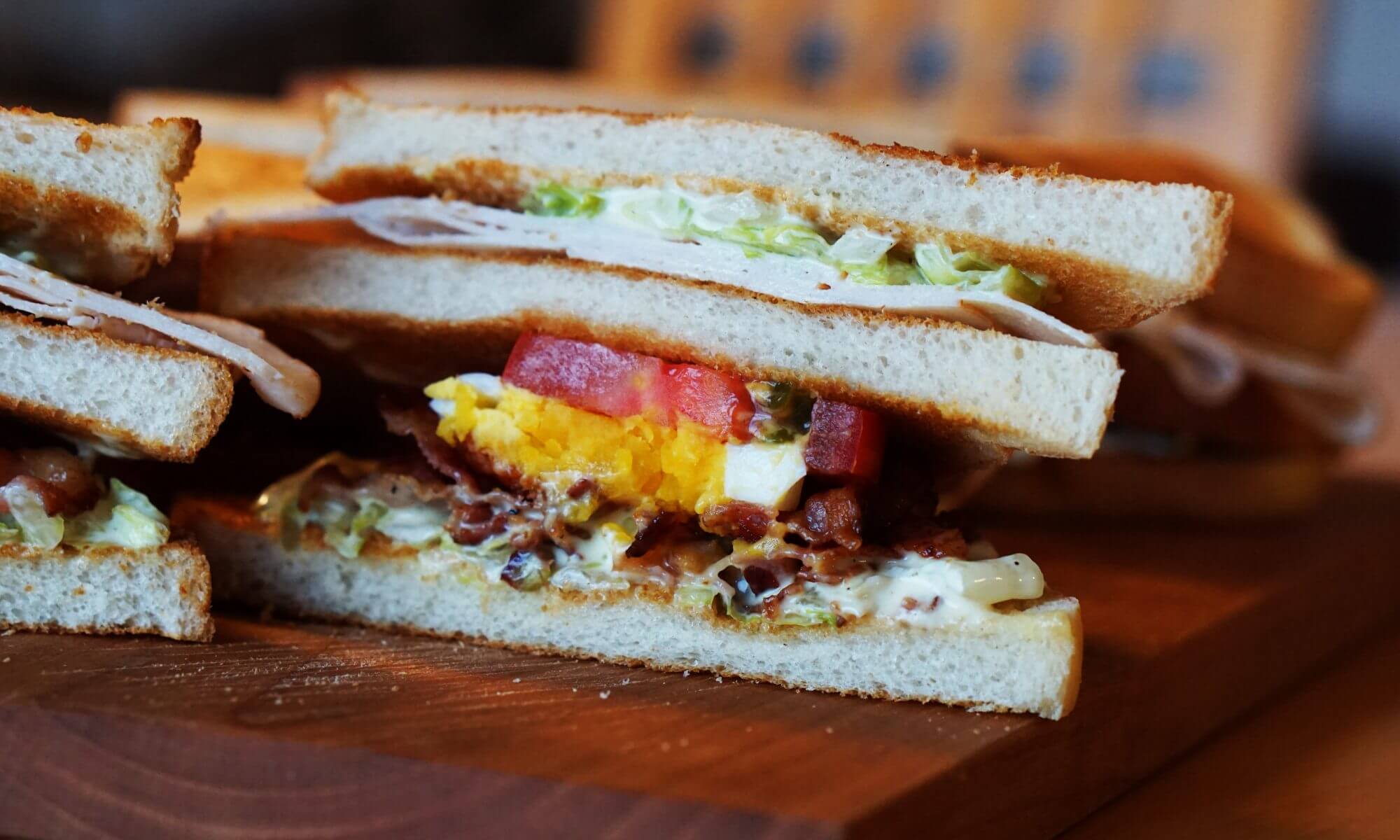 Extreme close-up of layered sandwich ingredients showing bread, egg, tomato, bacon, lettuce and sauce on a dark wooden surface, warm rustic tones, appetizing mood.